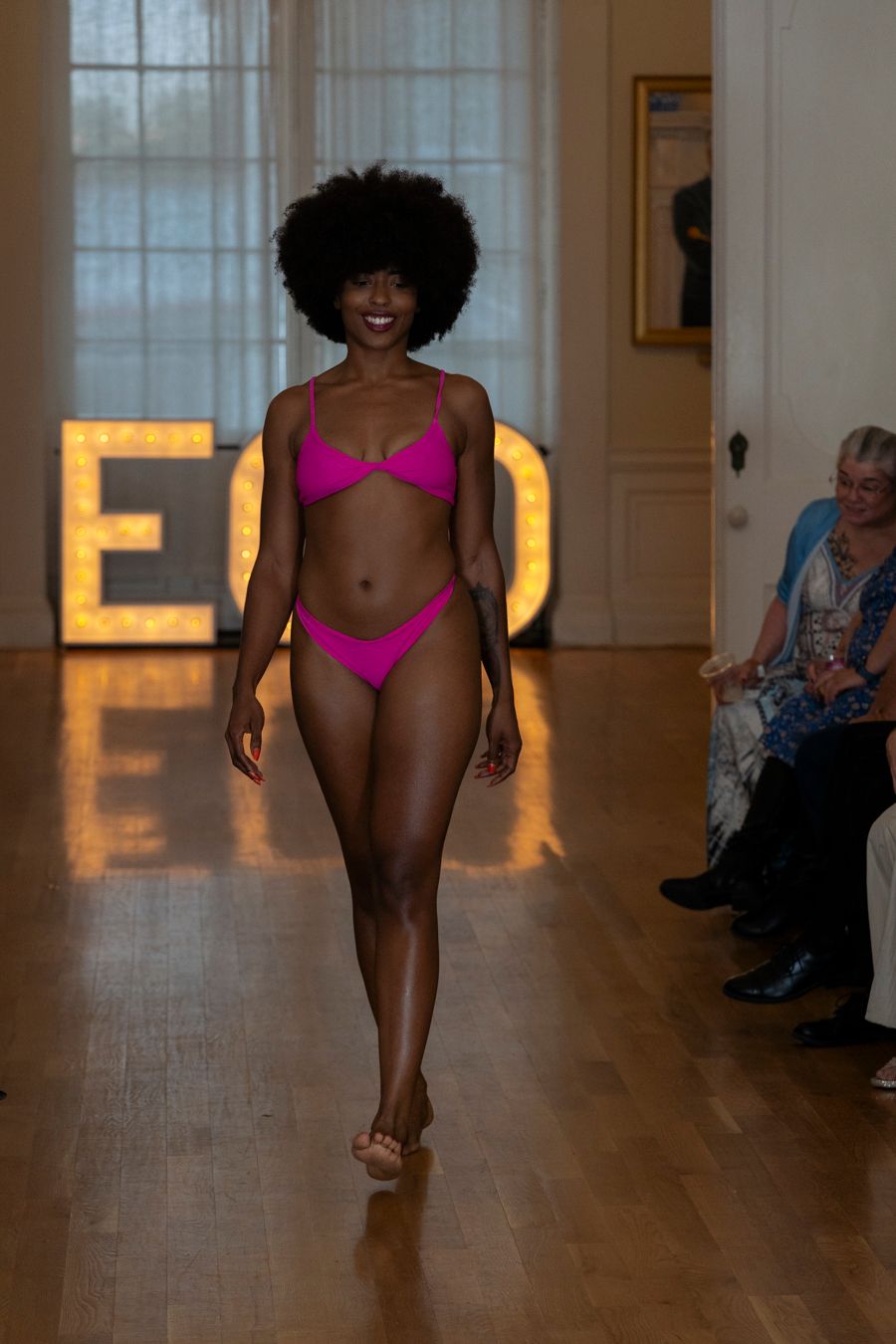 A fashion runway show featuring models walking in pink swimwear against a warm-lit hallway backdrop.