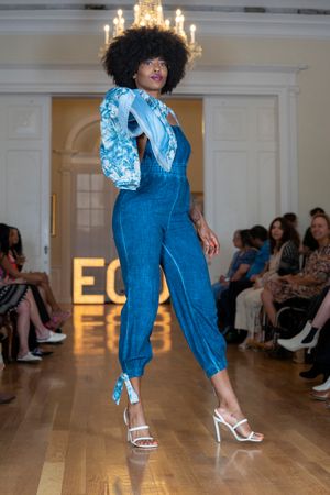 Model showcases denim jumpsuit with floral print jacket on runway under elegant chandelier lighting.