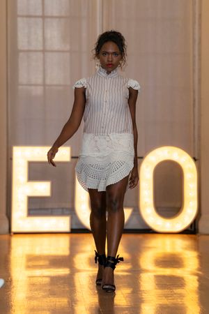Models walk the runway in white eyelet dresses and black shoes against illuminated letters during a fashion show.