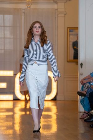 Model walks runway in striped white outfit with ruffled sleeves against warm lighting.