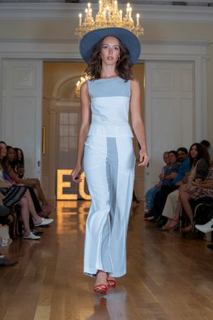 Fashion model walks runway in light blue sleeveless jumpsuit with wide legs and navy hat under elegant chandelier.