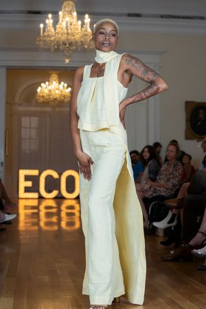 Model in flowing cream-colored ensemble walks runway under crystal chandeliers with illuminated ECO sign visible.