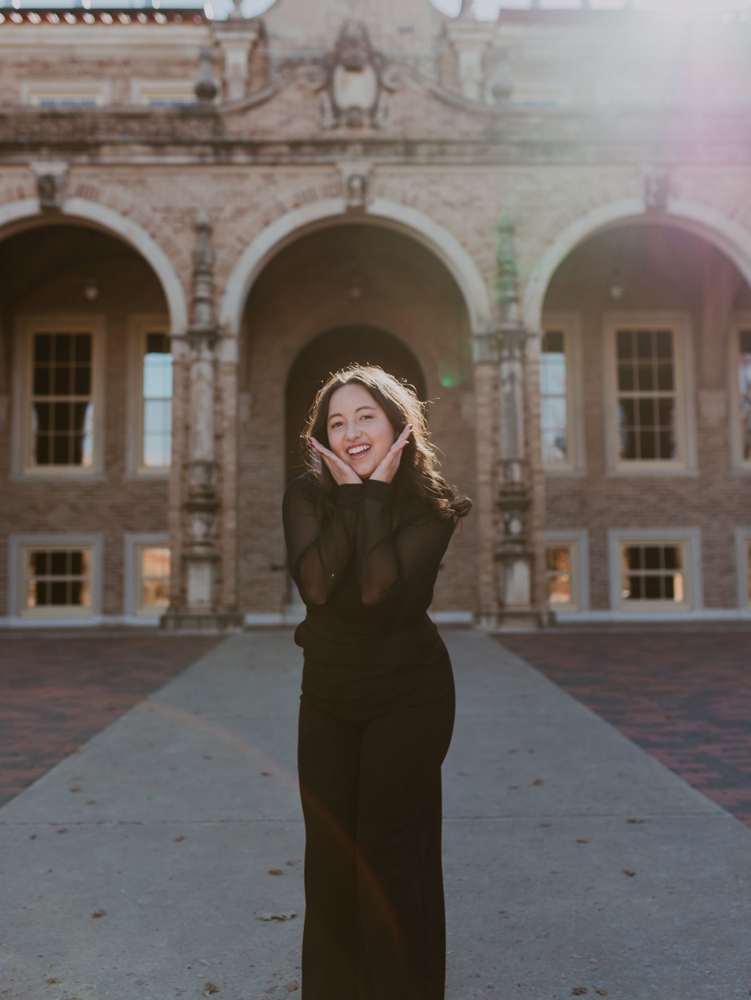 Smiling young woman in black outfit posing in front of historic brick building with arched windows.