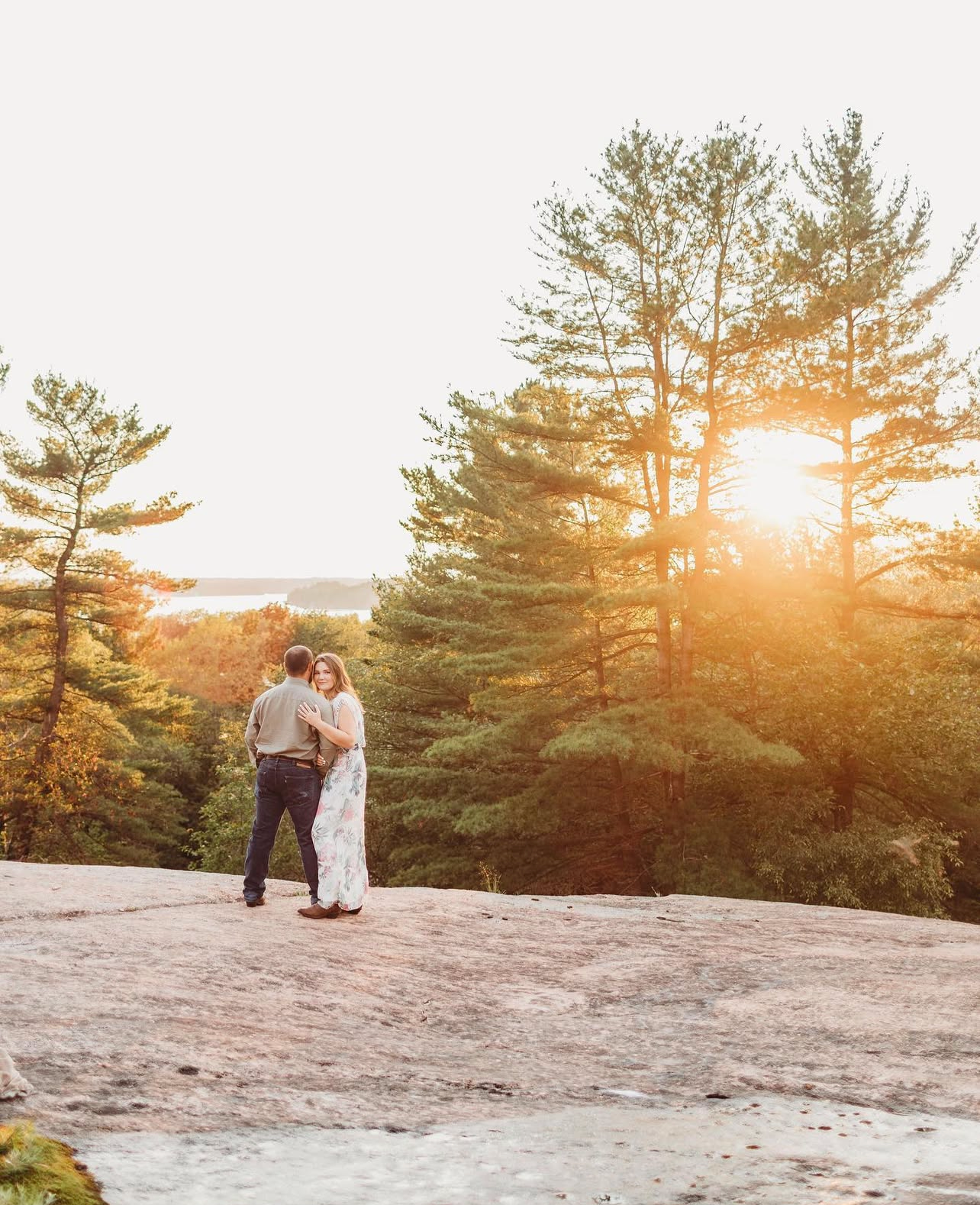 A couple shares a romantic moment on a rocky cliff at sunset with tall pine trees silhouetted against the golden sky.