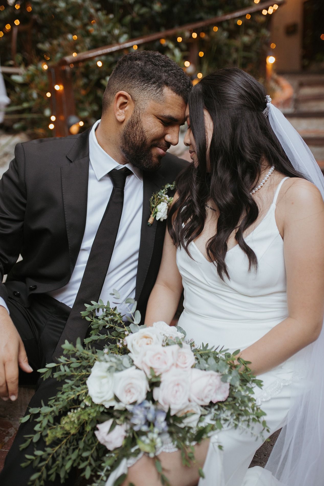 A romantic wedding couple sits close together with a lush bouquet of white flowers and greenery in an intimate setting.