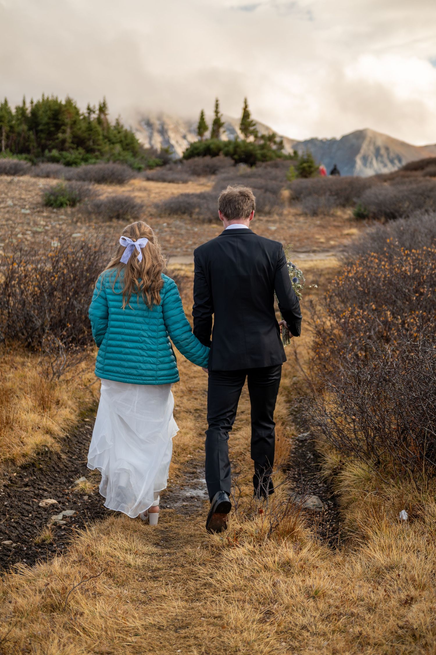 A bride and groom walk hand in hand in a grassy, rocky meadow. Dramatic clouds and mountain peaks in the background. She wears a teal coat.