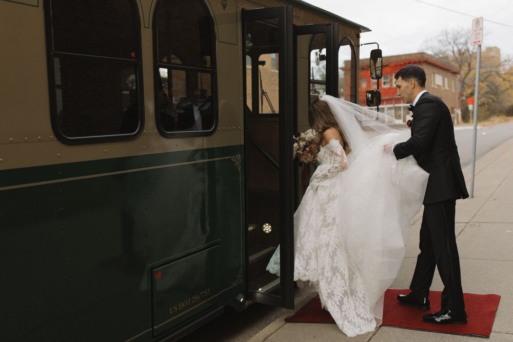 A bride in a flowing white dress is being helped onto a vintage train car by someone in dark clothing.