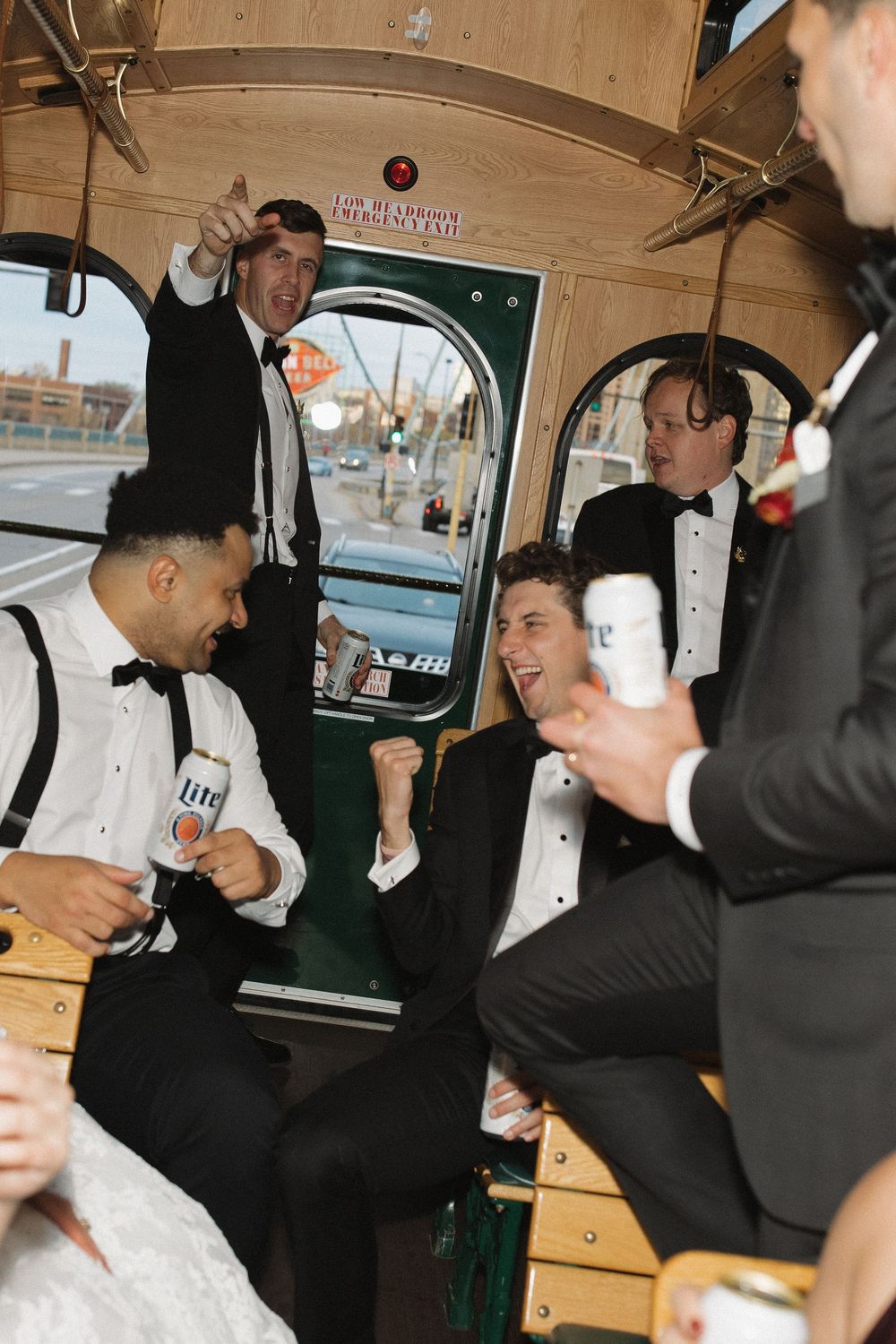 A group in formal tuxedos and suits share drinks and laughs while riding in a vintage vehicle interior.