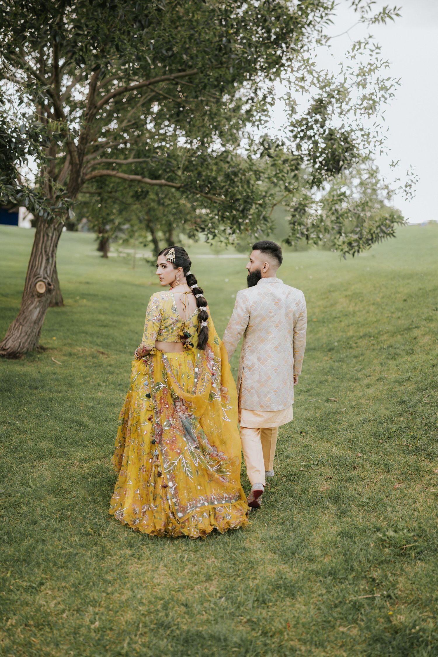 A couple in traditional South Asian wedding attire walks through a green garden with trees on a cloudy day.