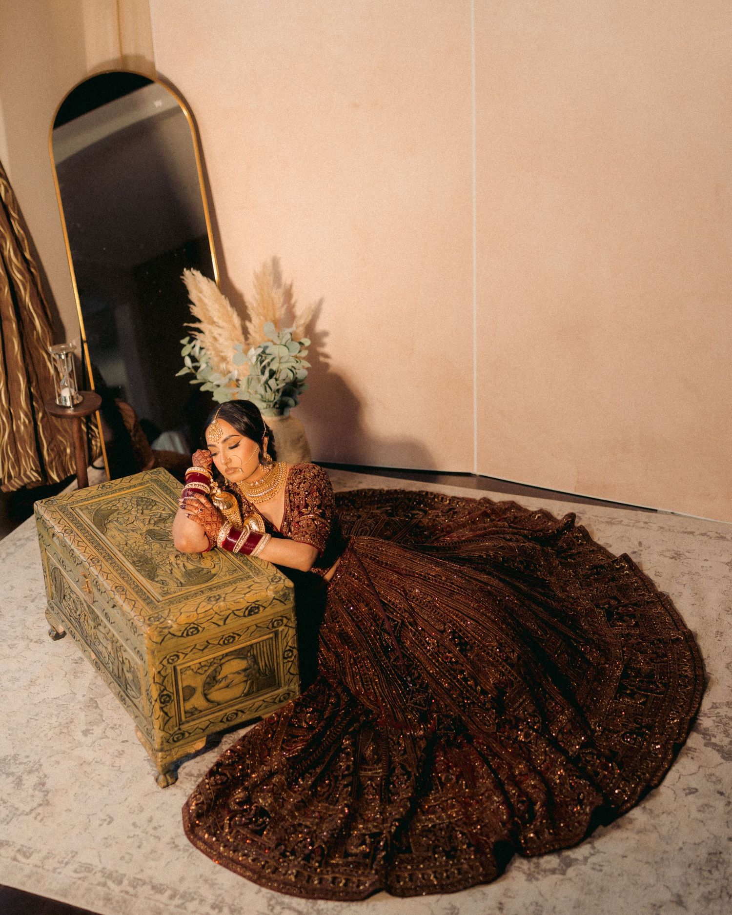 A model in an ornate brown lehenga poses elegantly on the floor against a light-colored wall in a fashion photoshoot.