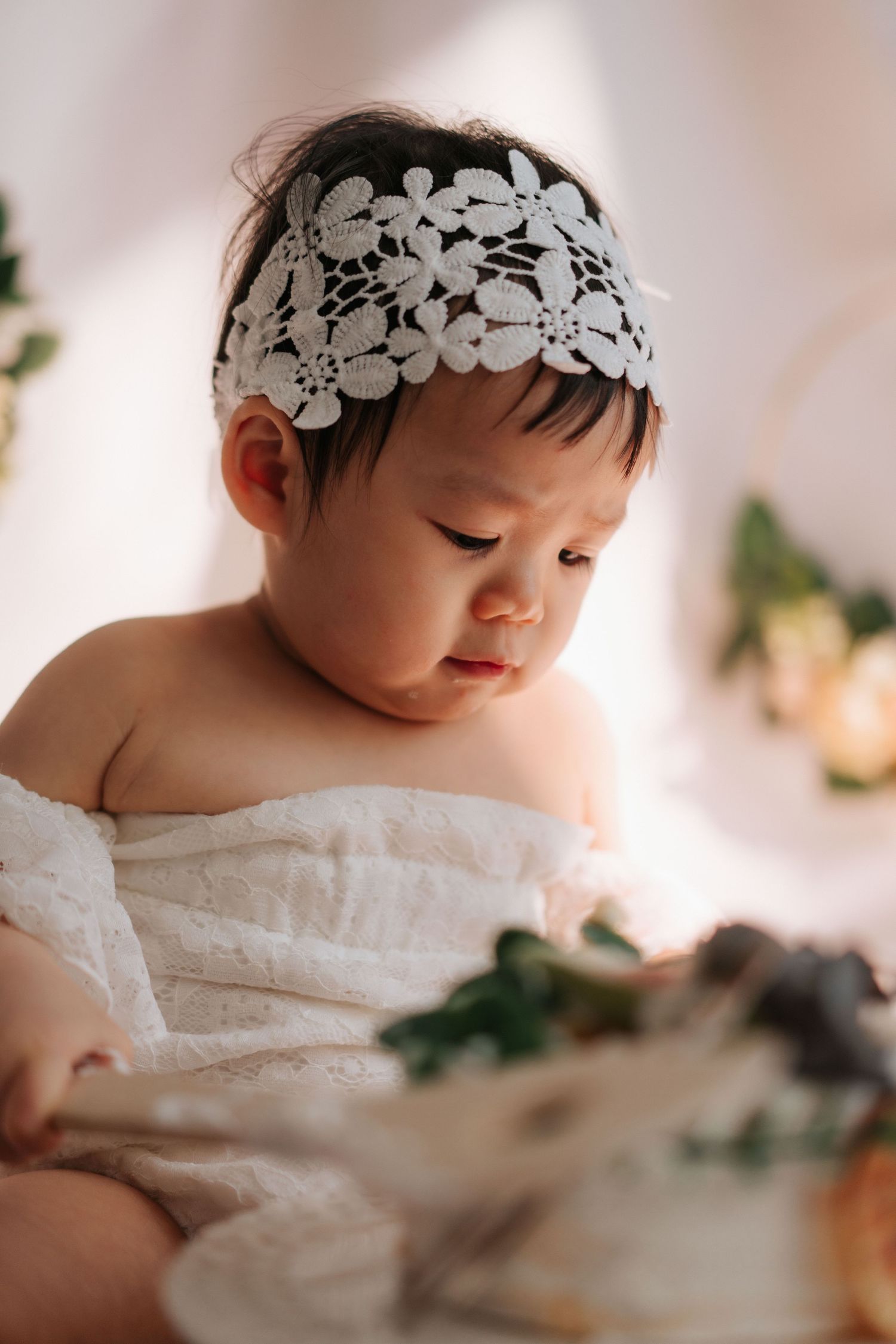 A baby wearing a delicate white floral headband and white dress gazes down in a soft, dreamy portrait series.