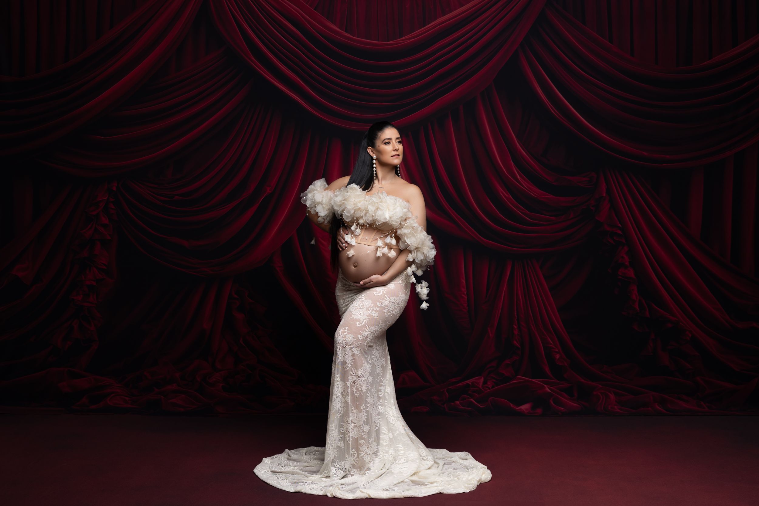 A bride in a white lace gown poses elegantly against deep red velvet theater curtains during a wedding photoshoot.