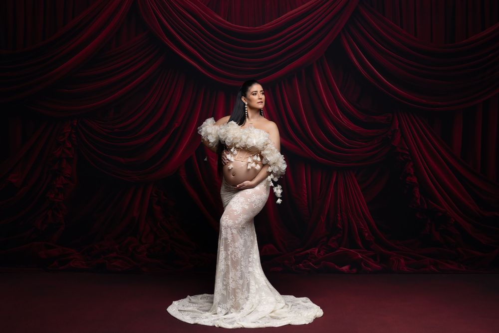 A bride in a white lace gown poses elegantly against deep red velvet theater curtains during a wedding photoshoot.