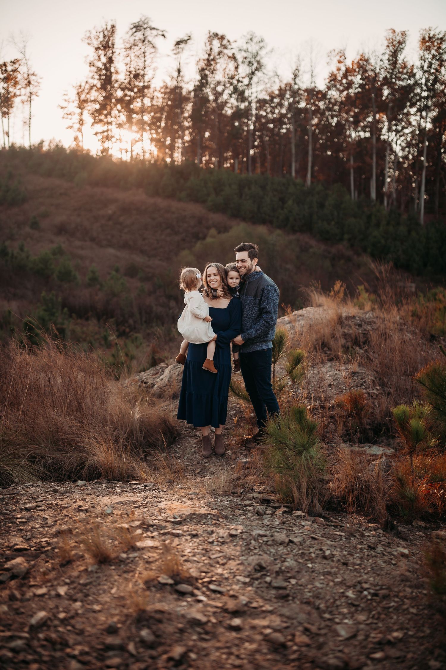 A family shares a tender moment on a rocky hillside trail during golden hour sunset.