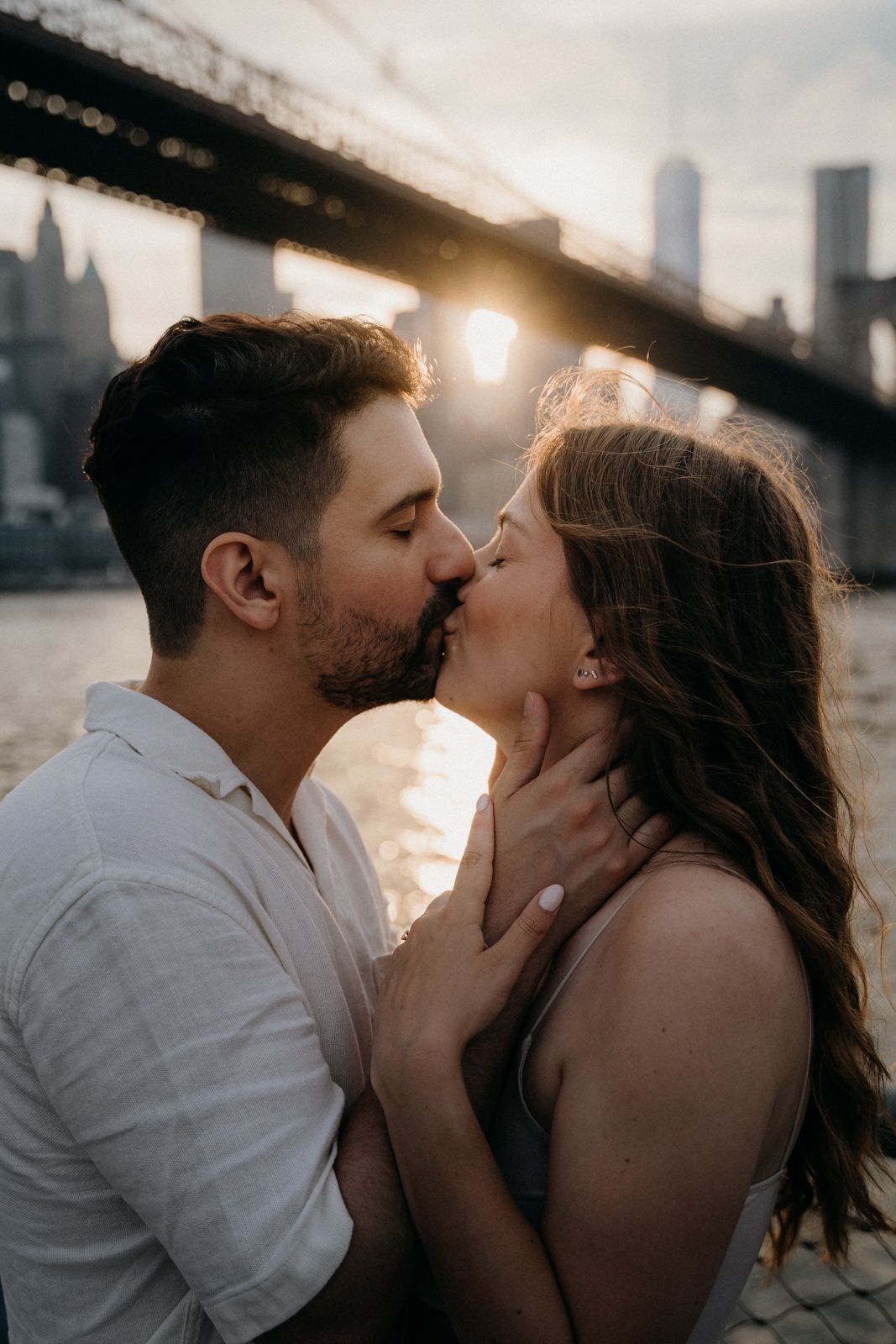 Romantic sunset kiss beneath Manhattan Bridge with city skyline in background.