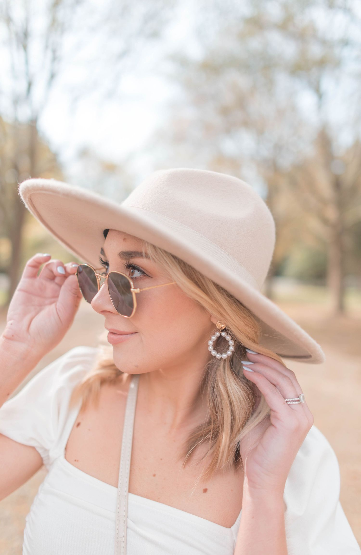 Woman in beige wide-brim hat and gold hoop earrings adjusting rose-tinted sunglasses outdoors in a park.