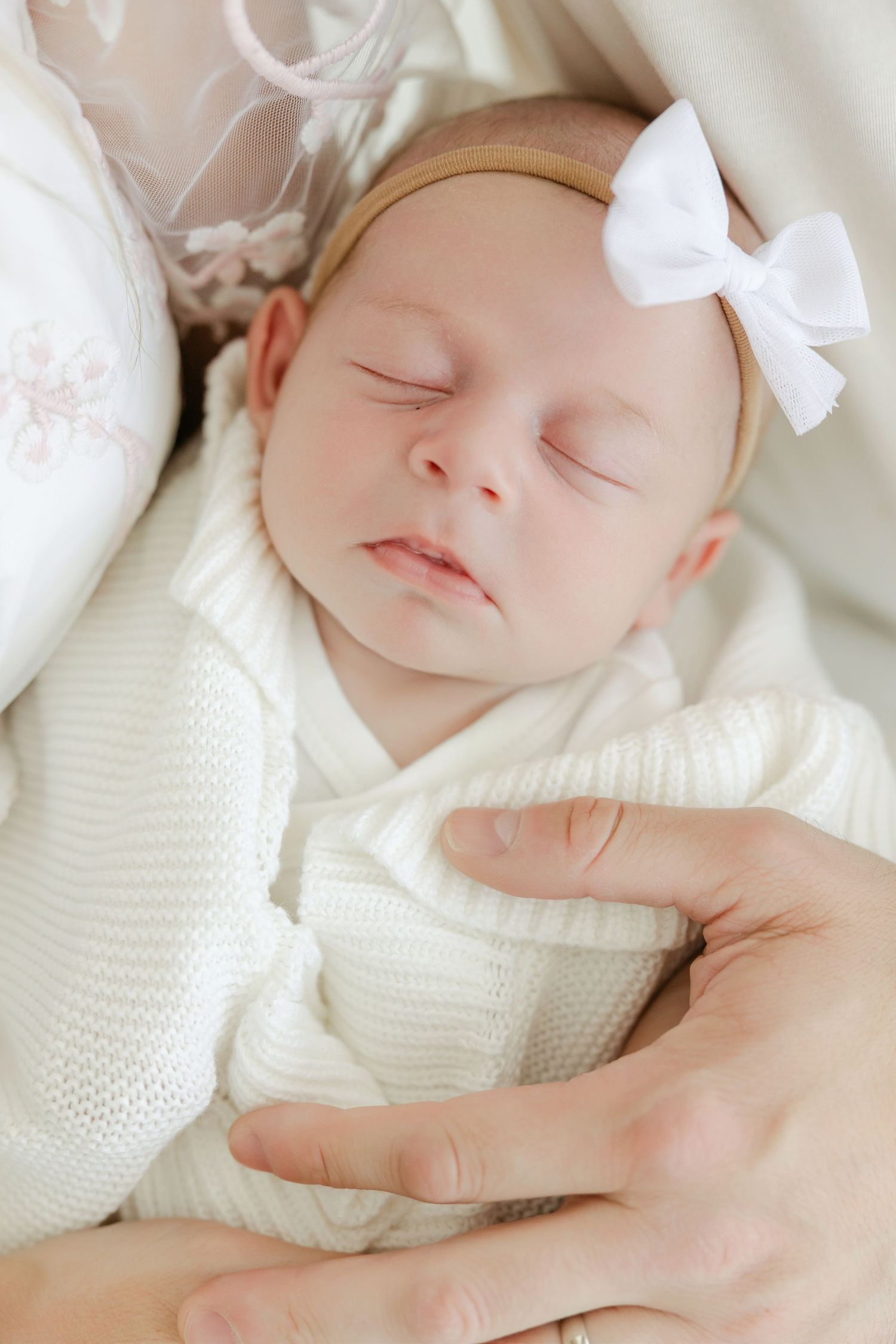 A sleeping newborn baby wearing a white bow and knit sweater lies peacefully on soft white bedding. Connecticut Newborn Studio.
