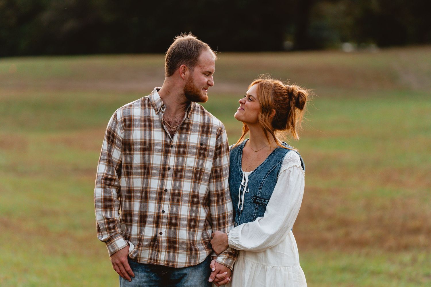 A couple holding hands and gazing at each other while standing in a grassy autumn field.