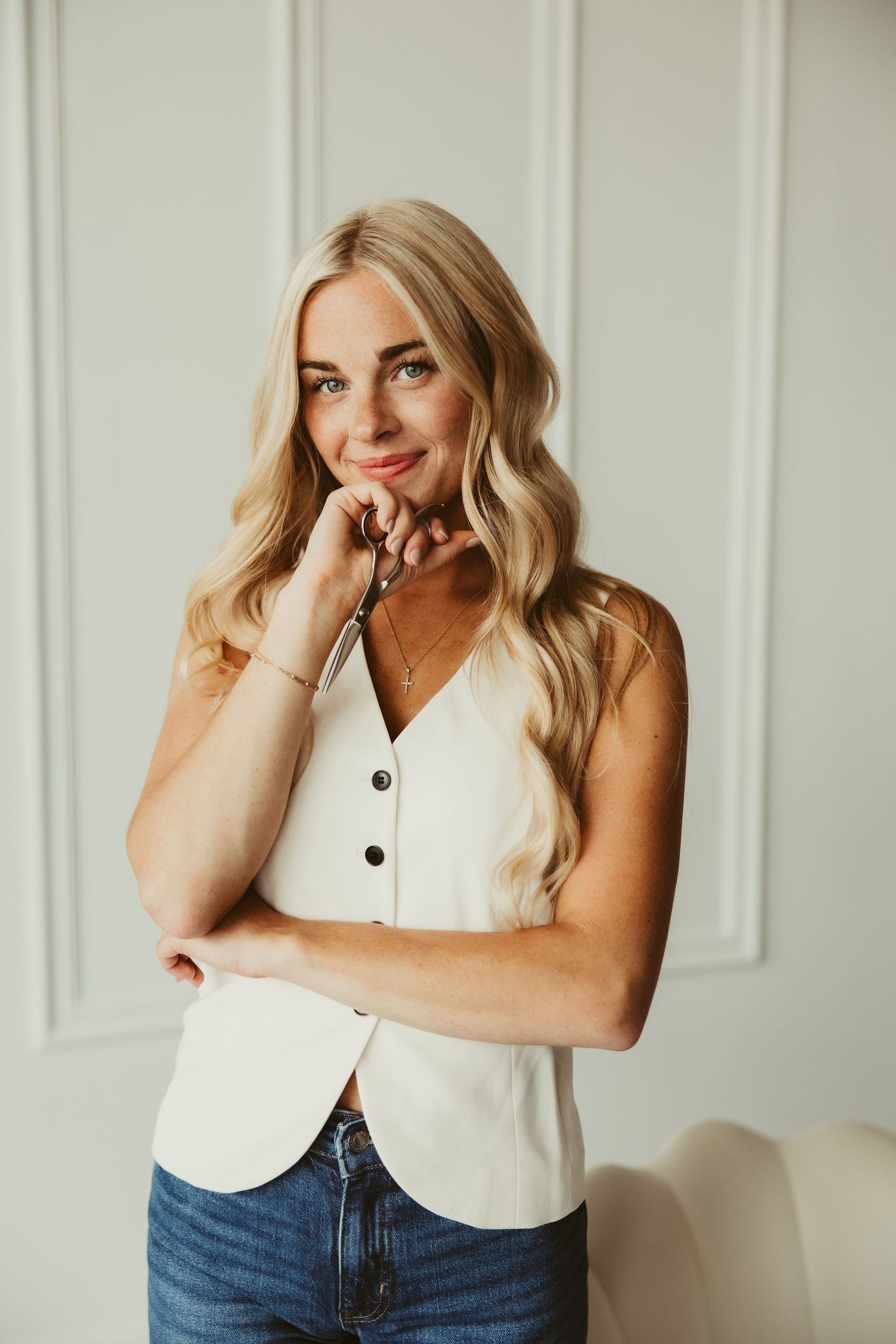 Smiling blonde woman in white sleeveless top posing against white wall.