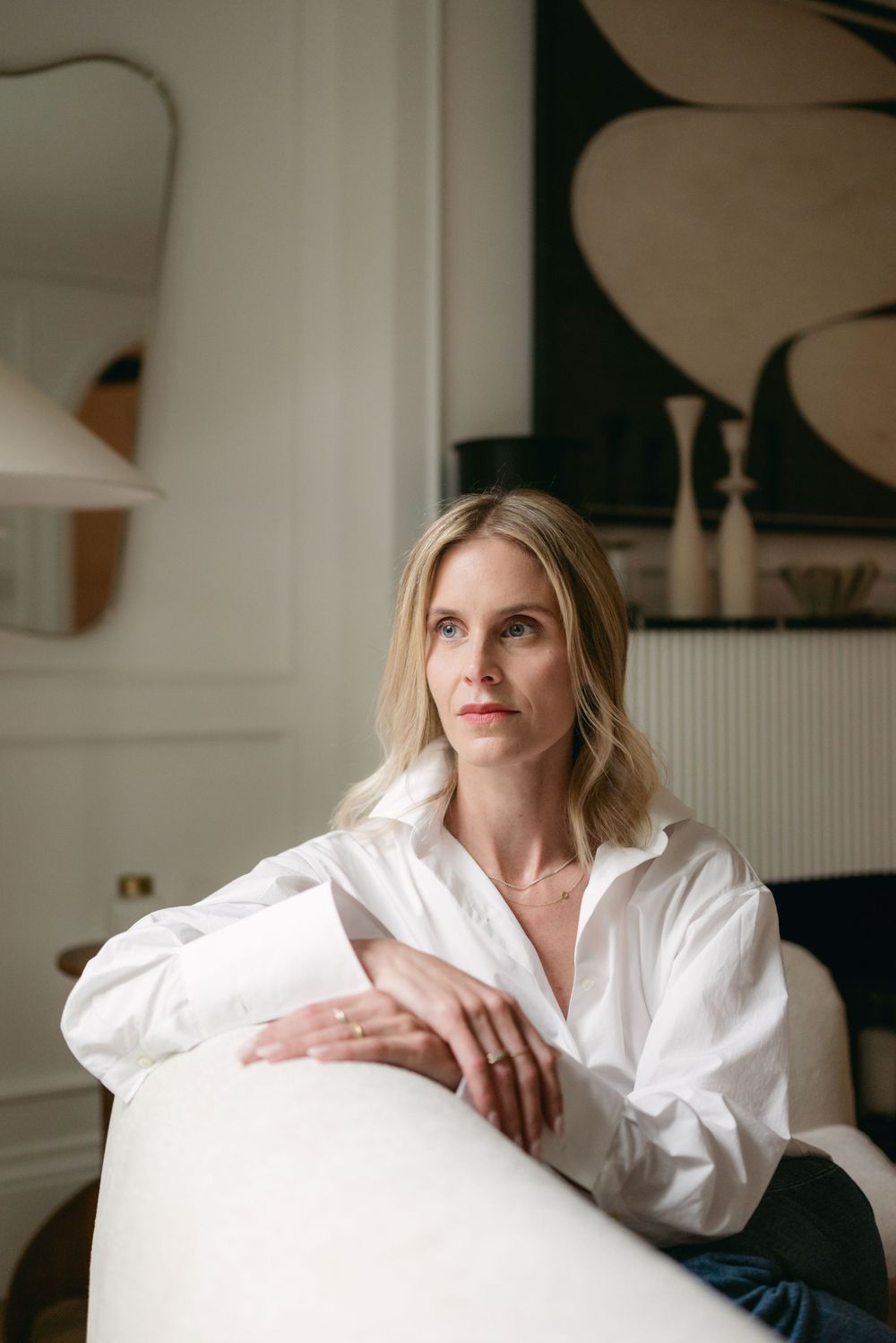 A person in a crisp white button-down shirt sits pensively on a white chair in a minimalist interior setting.