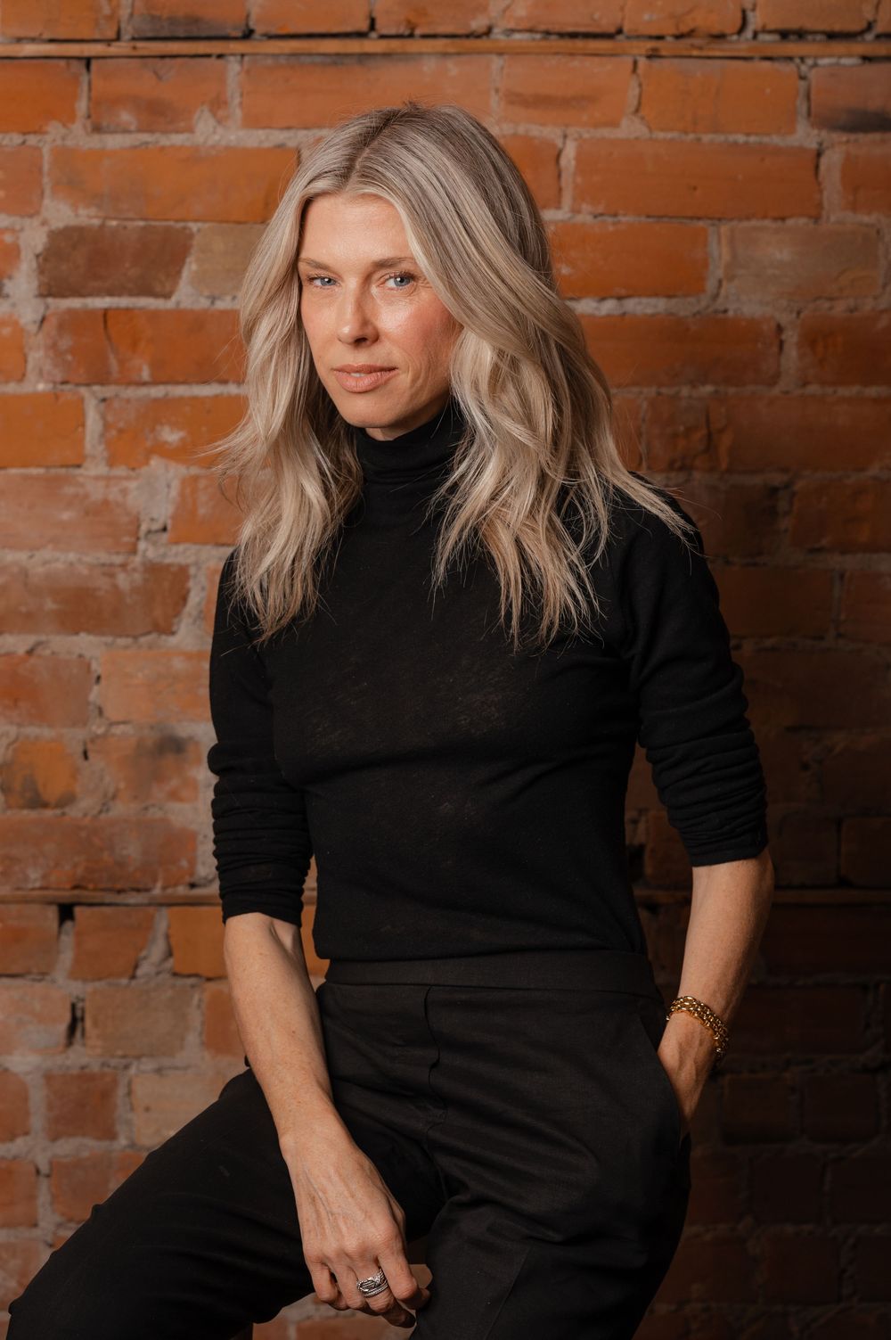 A series of four artistic portraits against a rustic brick wall background showing someone in a black turtleneck sweater.