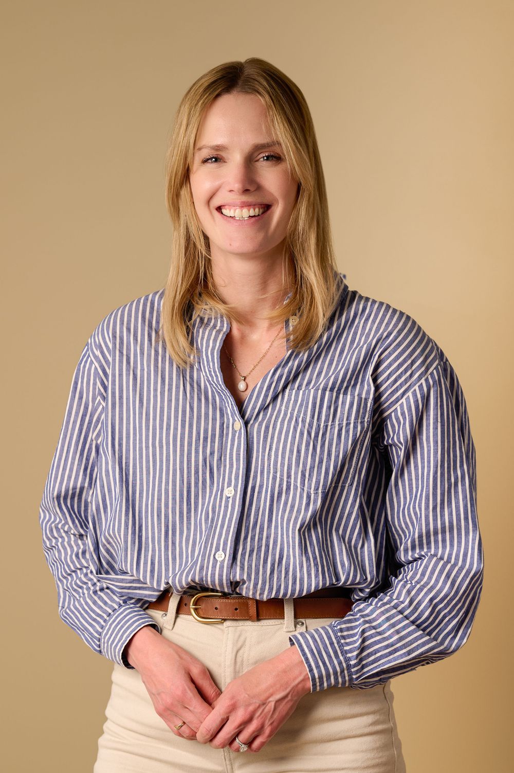 Professional portrait of someone wearing a blue striped button-down shirt and khaki pants against a beige background.