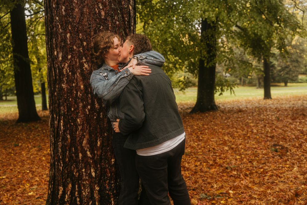 A couple embraces in a park surrounded by autumn leaves and tall trees on a romantic fall day.