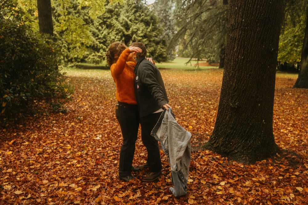 Two people share an intimate moment among fallen leaves in an autumn forest setting.