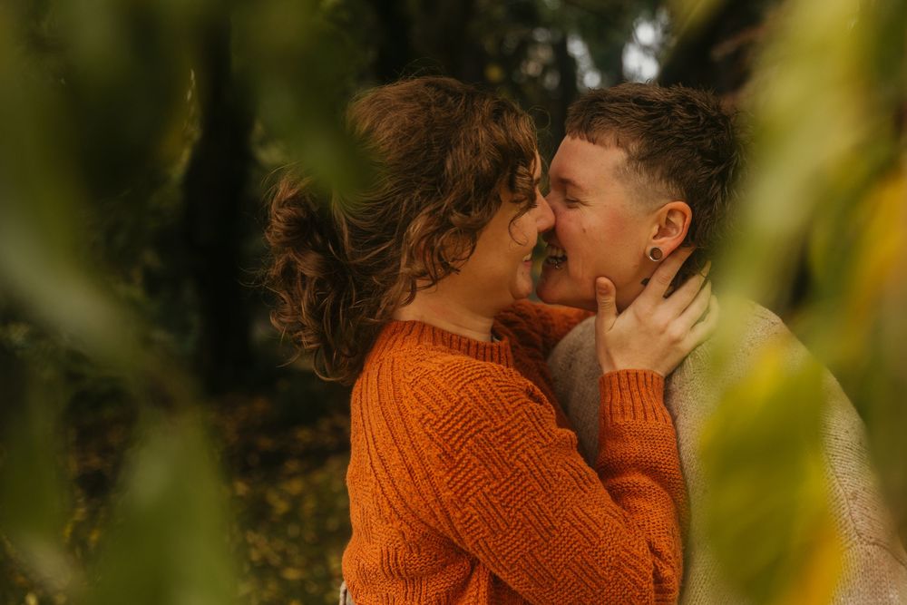 Couple in autumn setting wearing orange sweater embracing among blurred foliage.