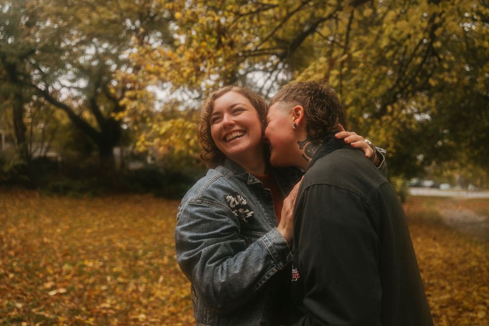 Two people laugh and embrace joyfully in a park filled with fall colors.