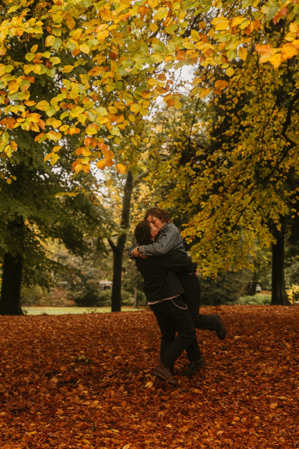 Two people share an intimate moment among fallen leaves and autumn trees in a park.