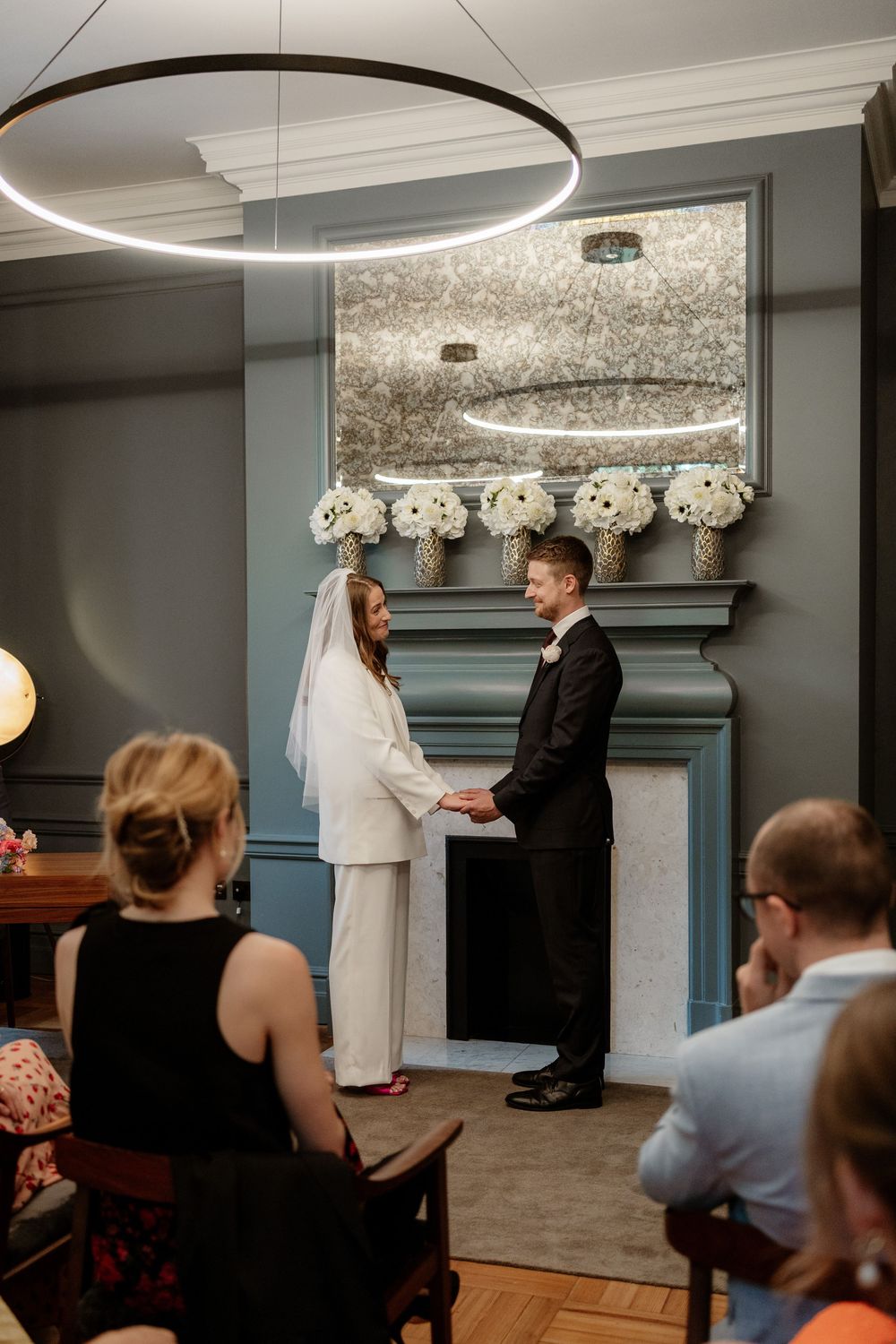 A modern wedding ceremony in an elegant room with a circular light fixture and floral decorations above a grey fireplace mantel.