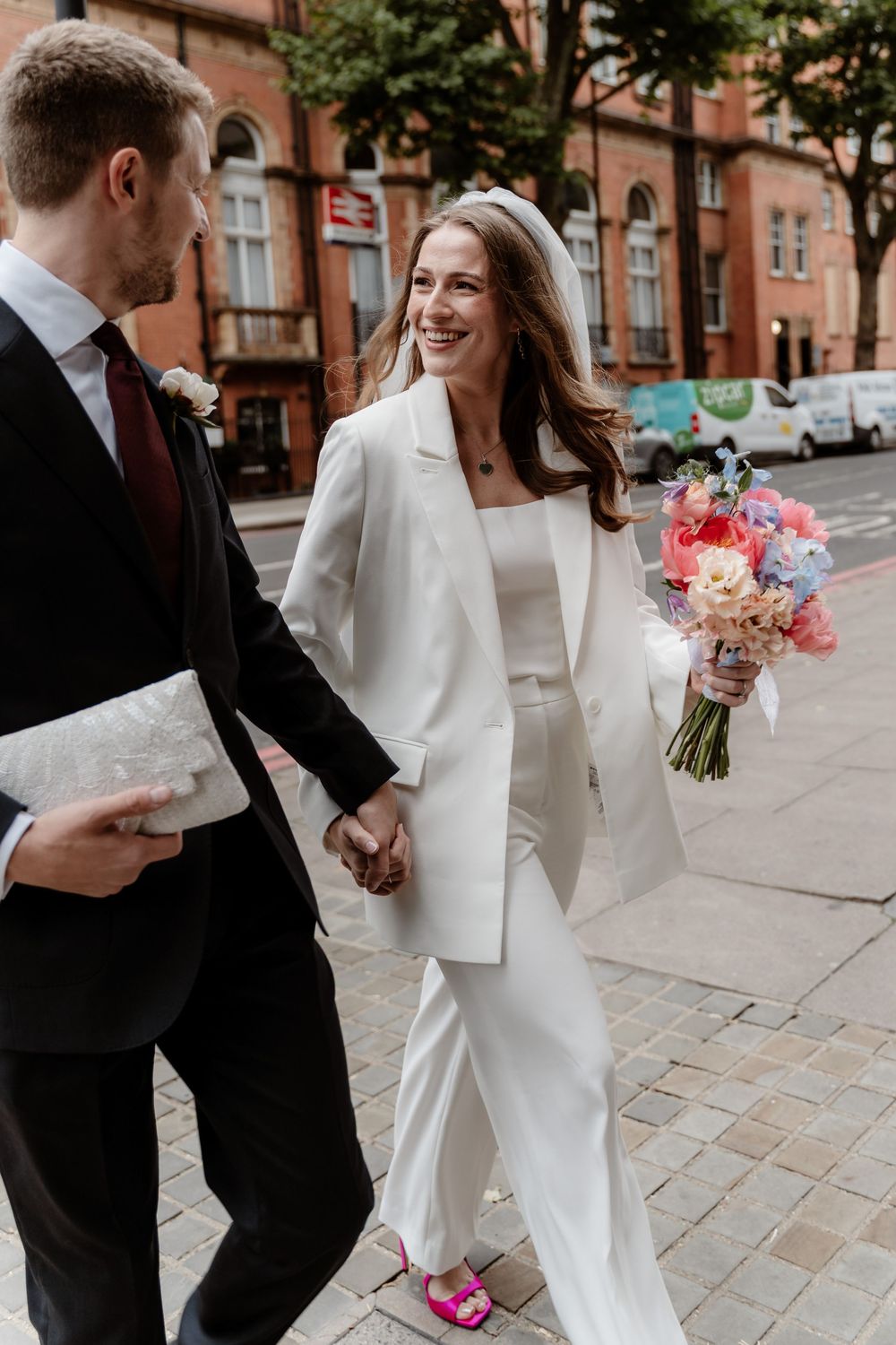 A couple in wedding attire walks together on a brick sidewalk holding hands and a pink floral bouquet.