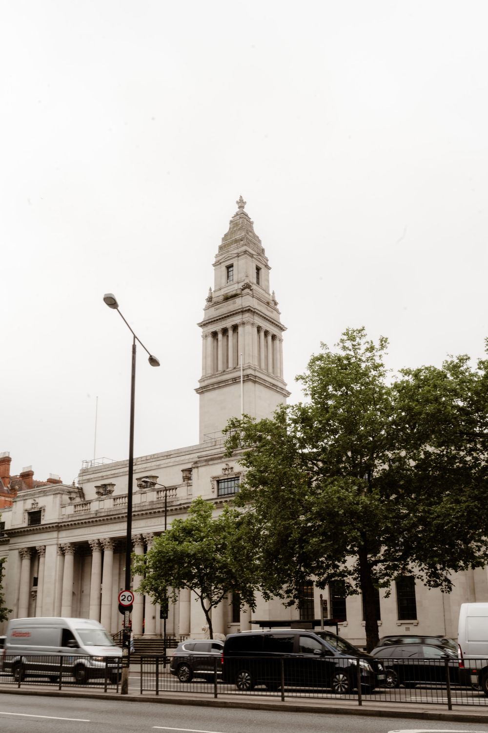 Historic white stone building with classical architecture and towers stands prominently along a tree-lined street in Cardiff.