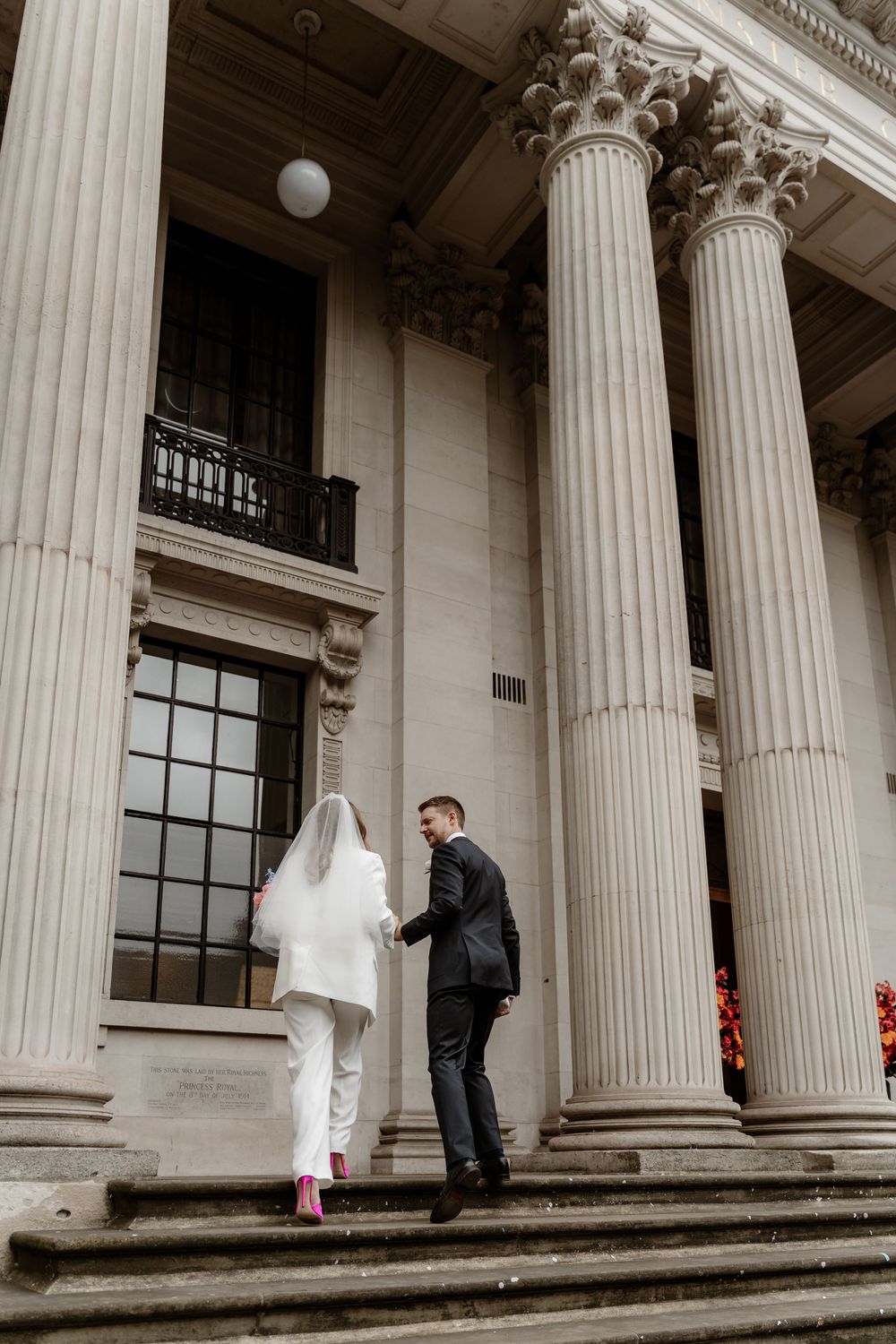 A couple in wedding attire stands between classical white stone columns on the steps of a grand building.