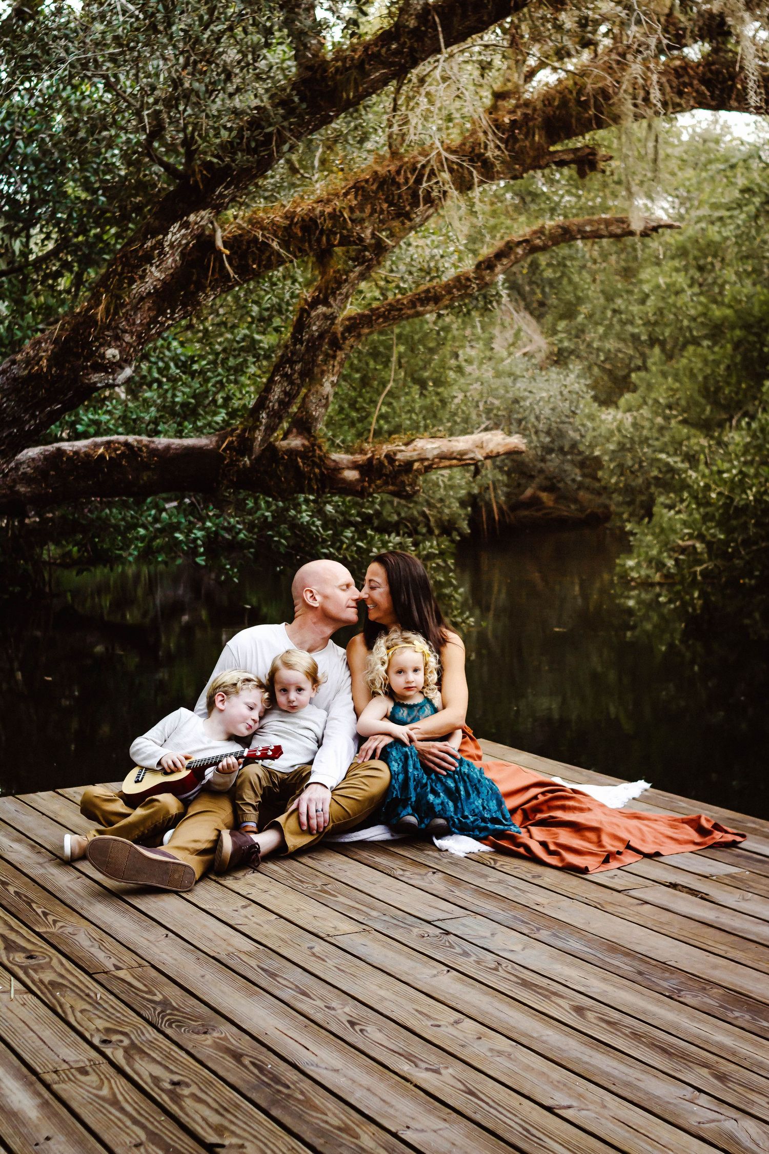 Family lying together on a wooden dock over water surrounded by lush greenery.