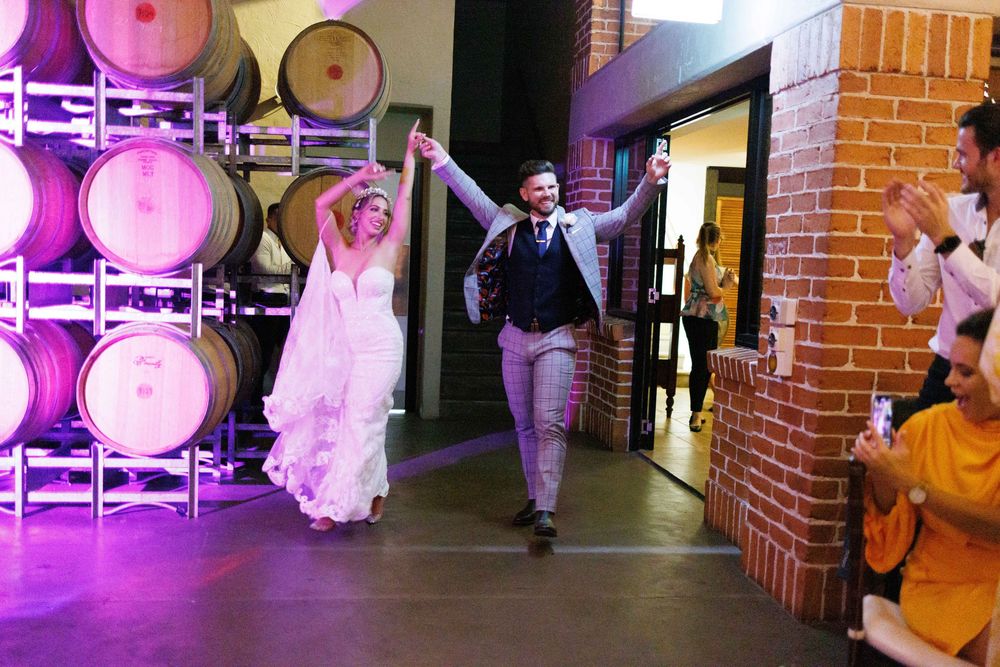 Bride and groom make a joyful grand entrance at a winery wedding reception surrounded by oak barrels.