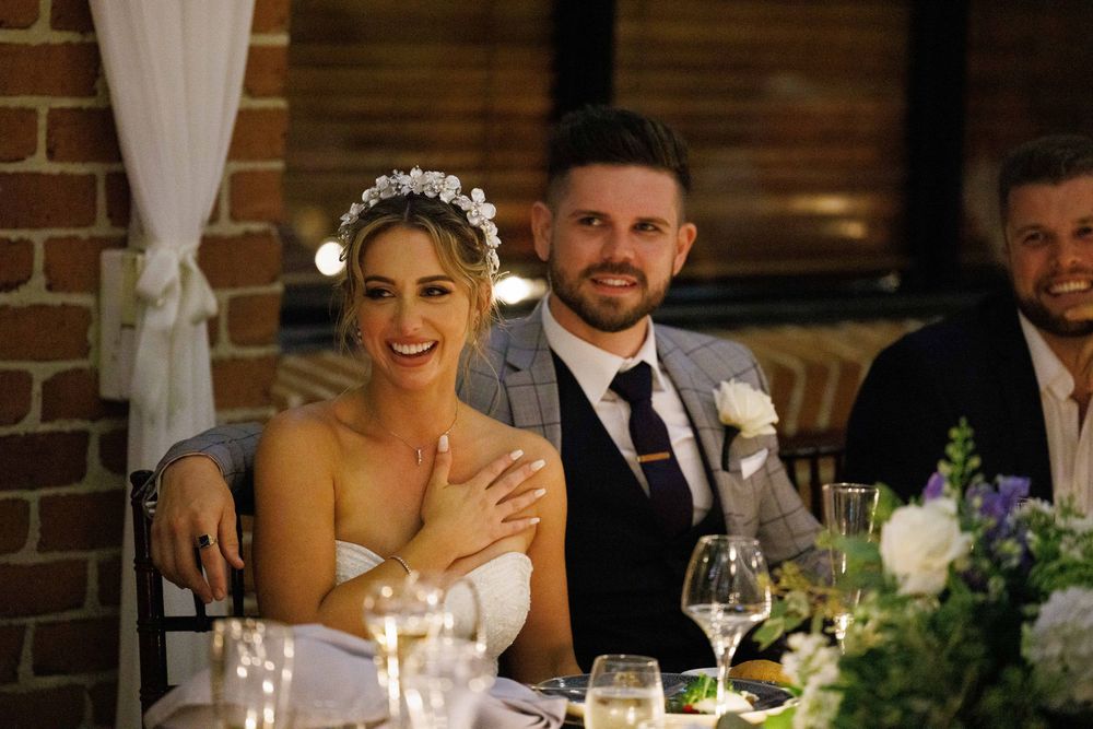 Bride wearing floral crown and groom in grey suit smile at wedding reception table.