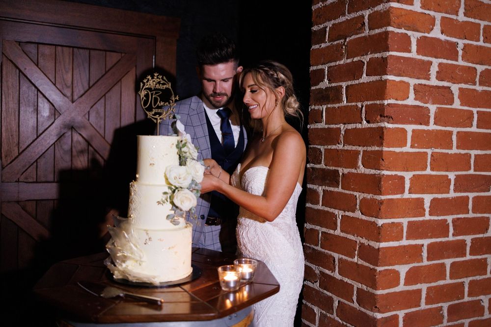 Bride and groom cut three-tier white wedding cake decorated with flowers by brick wall.