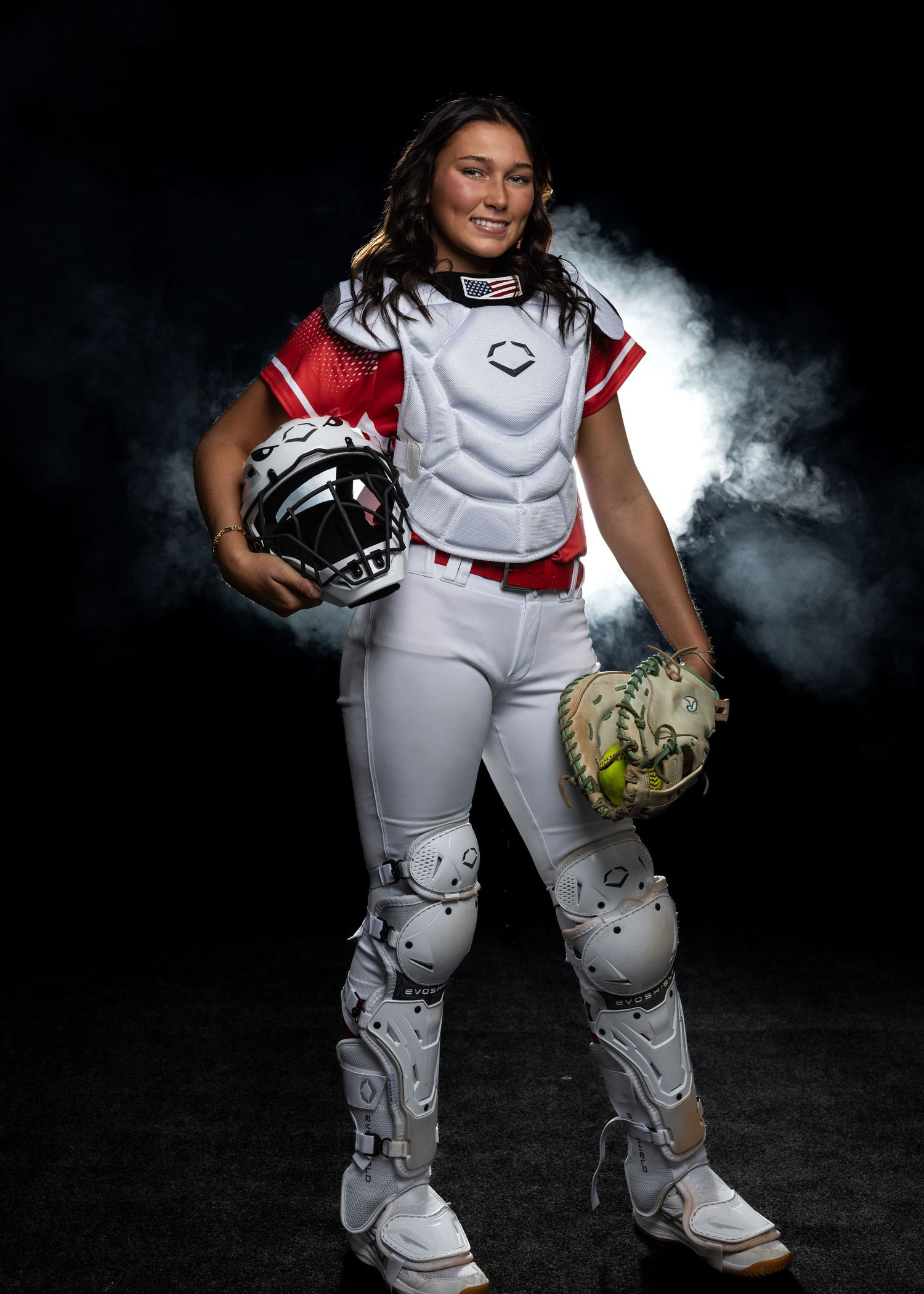 Softball player poses in white uniform with protective gear and helmet against dark background with atmospheric smoke effect.
