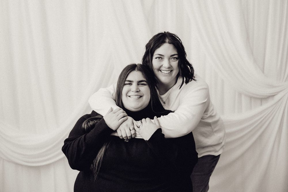 Two smiling women hugging in a black and white studio portrait with white curtain backdrop.