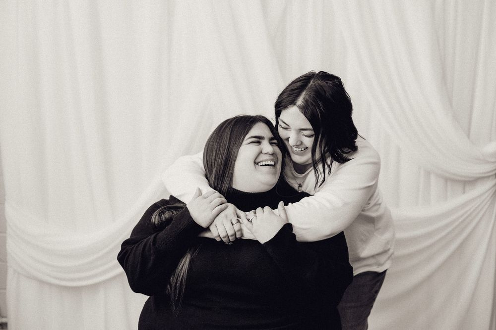 Two laughing women embracing playfully in a black and white studio portrait photo.