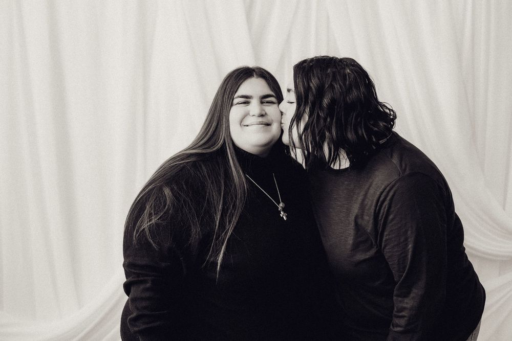 Two women in black and white studio portrait, one whispering in the other's ear while she smiles.