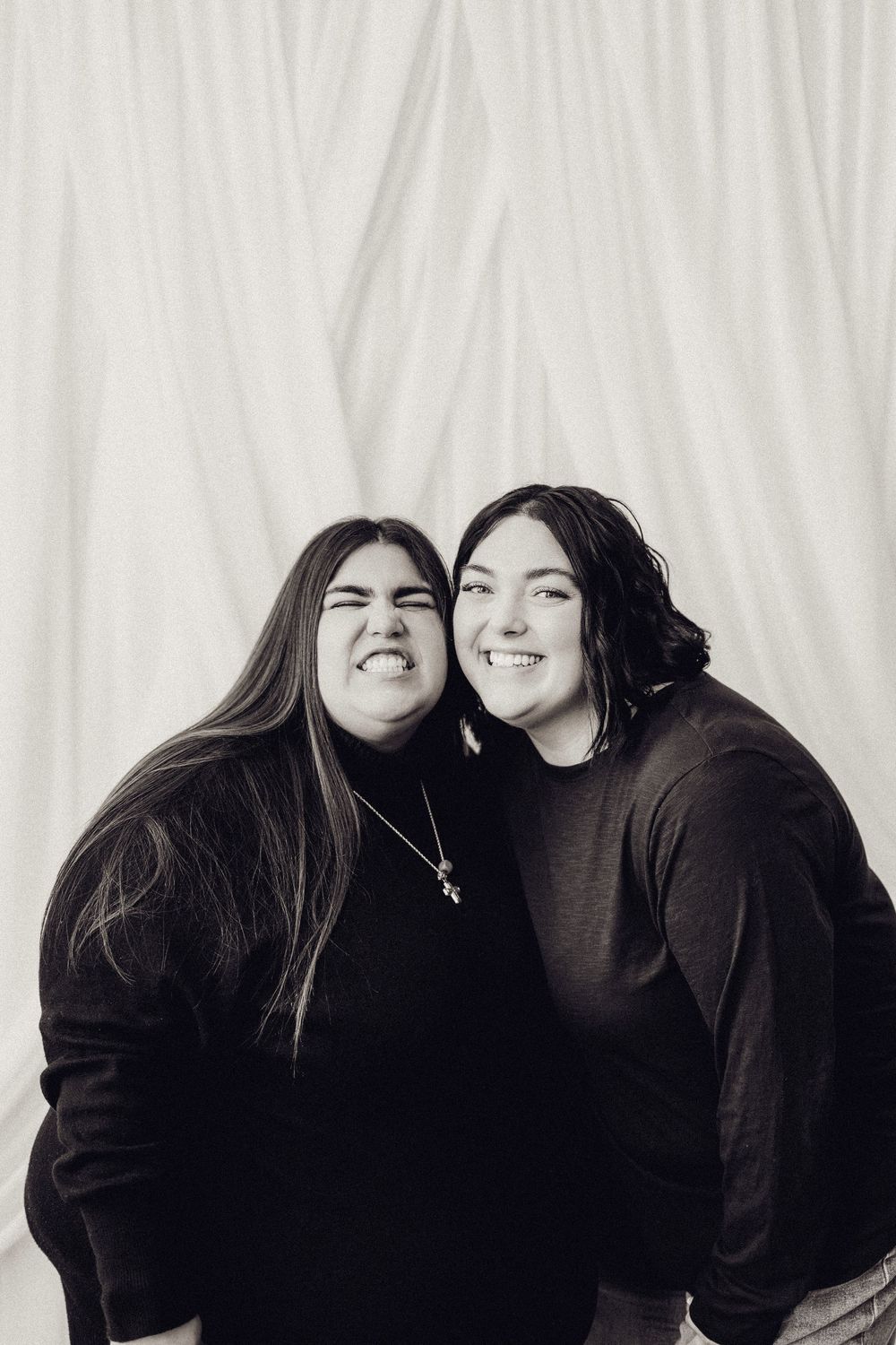 Two women smiling and making funny faces in a black and white studio portrait.