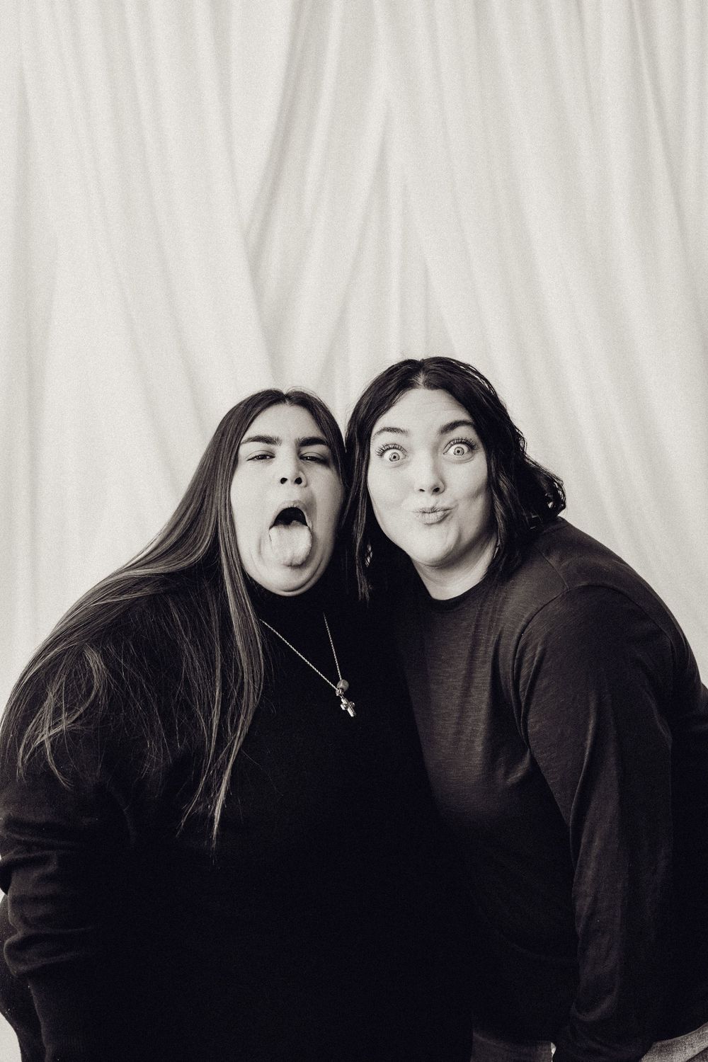 Two women making silly faces with tongues out in a black and white studio portrait.