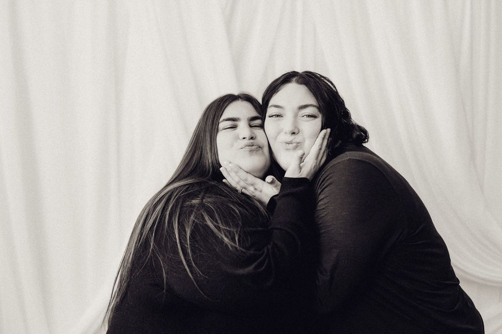 Two young women making kissing faces cheek to cheek in a black and white studio portrait.