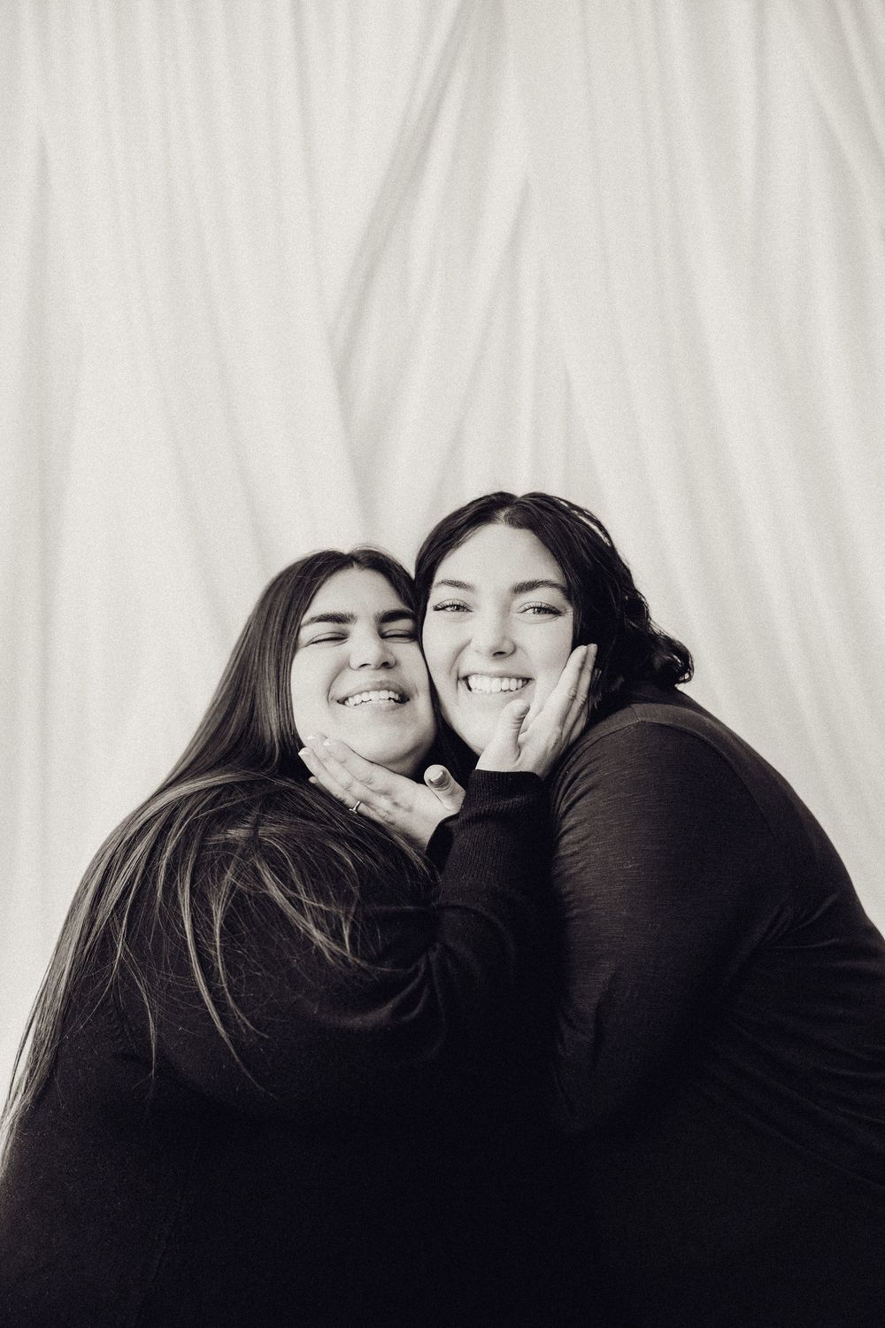 Two young women in black tops laugh and cuddle together in a black and white studio portrait.
