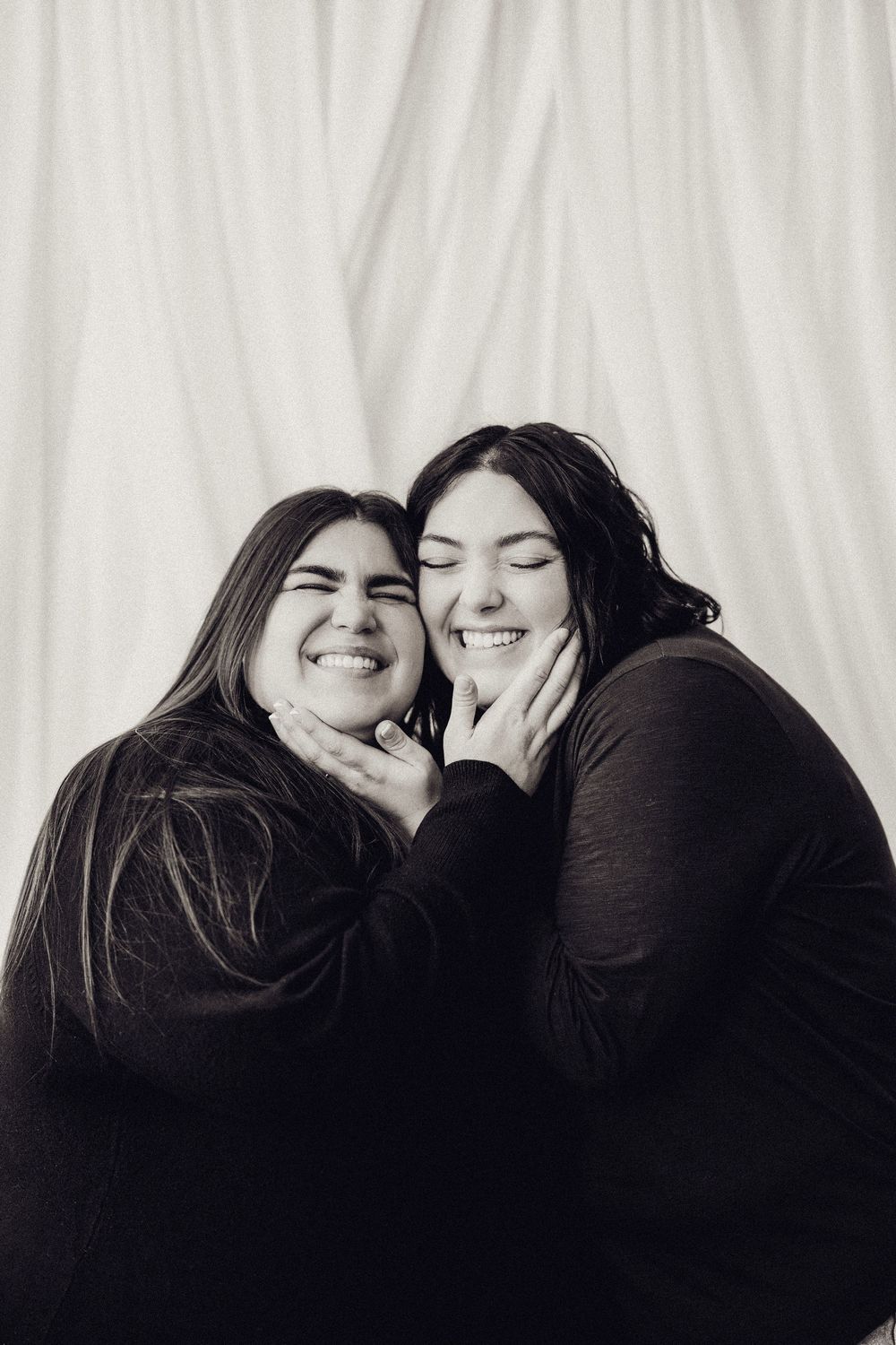 Two smiling young women in black outfits share a joyful moment in a black and white photo.