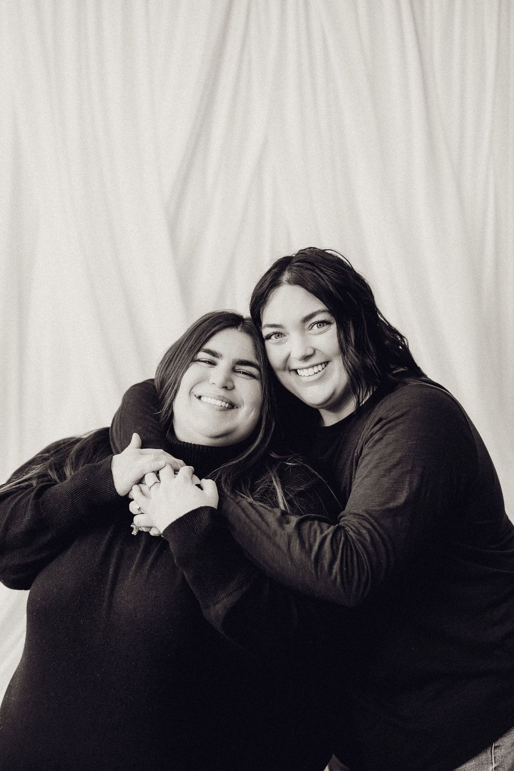 Two women in black tops embrace warmly, smiling in a black and white studio portrait.