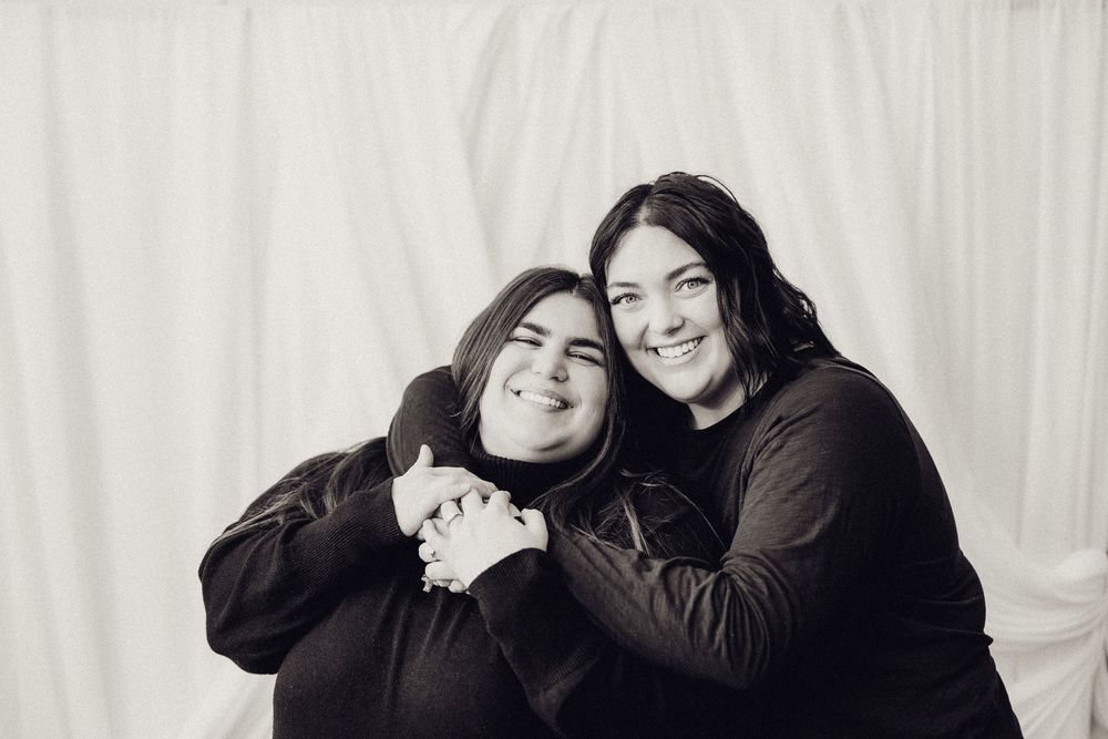 Two women in black clothing share a close hug in a black and white studio photo.